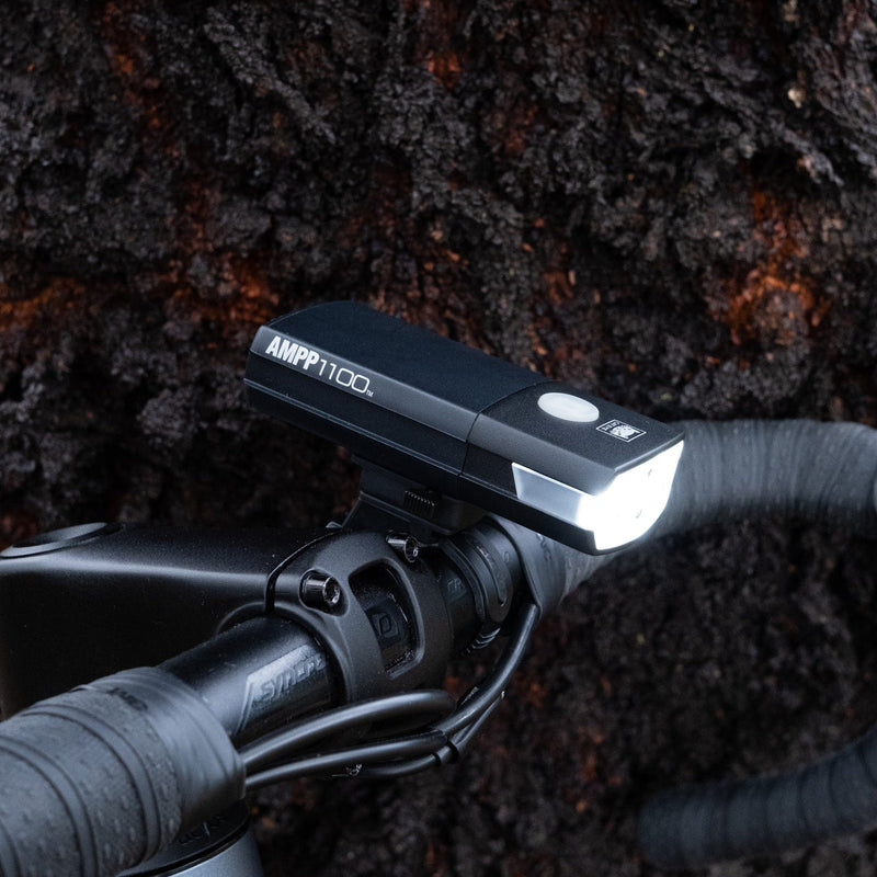 A black AMP 1100 bicycle handlebar light mounted on a drop handlebar, shown against a dark textured tree bark background.